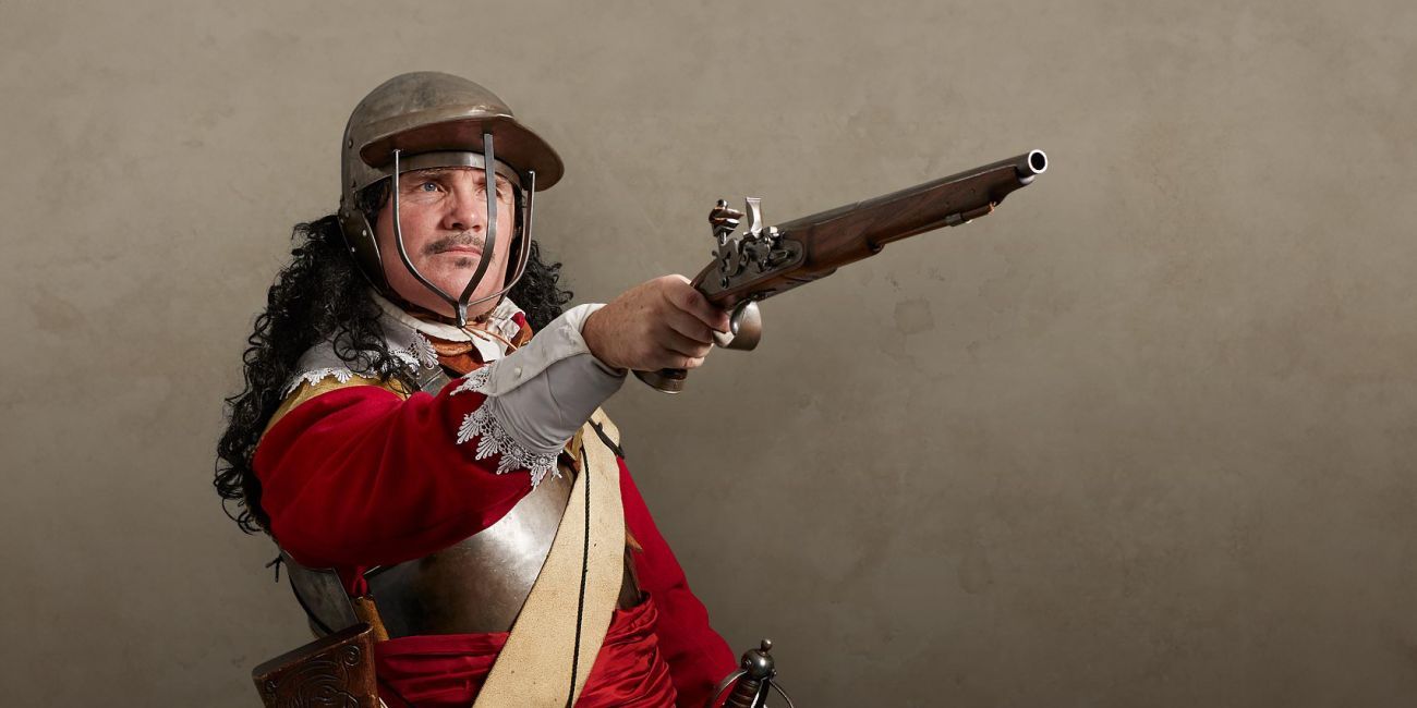 Royalist cavalier soldier dressed in a red tunic, buff coat, breastplate and a three-bar helmet, pointing a flintlock pistol
