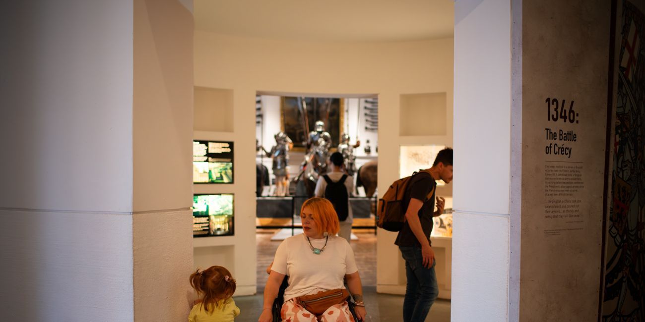 a visitor in a wheelchair navigates a gap in a gallery comfortably while passing a small child who leans against the wall