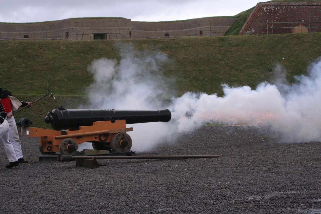 Large canon being fired by two men in period costumes with Fort Nelson in the background
