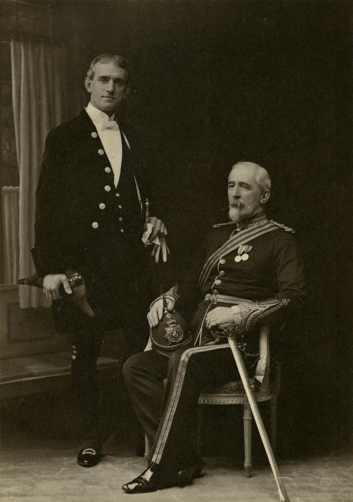 a man standing dressed in court dress beside a seated gentleman in uniform