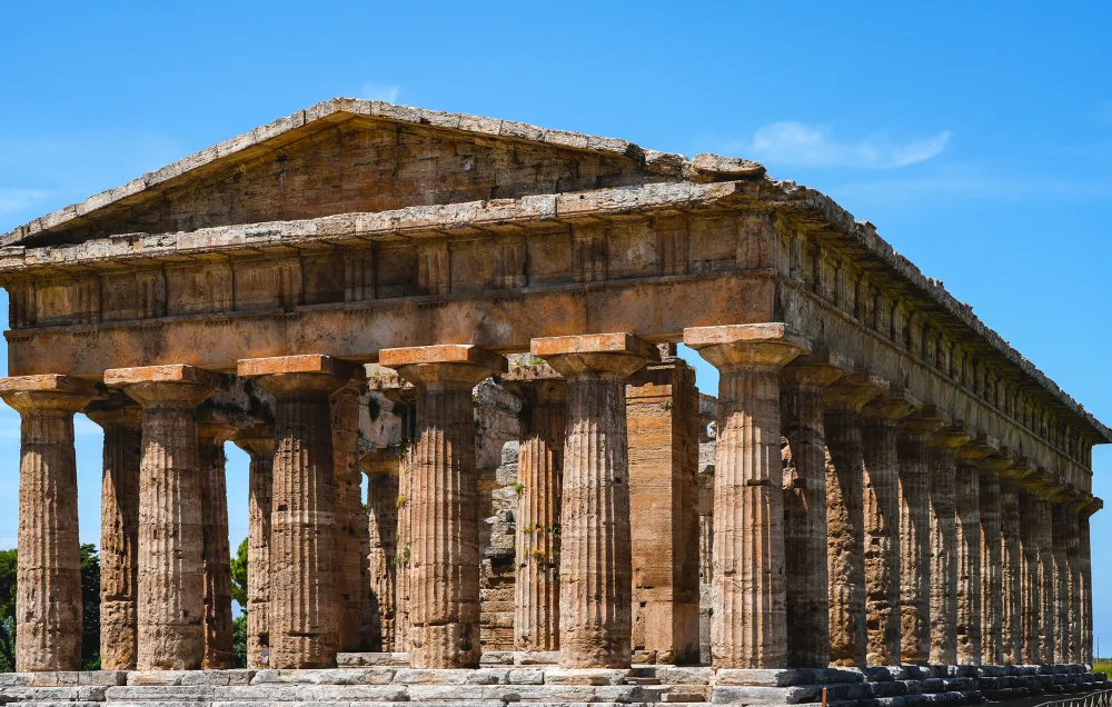 Ancient Greek Doric temple ruins with fluted columns and a triangular pediment under a bright blue sky. Temple of Poseidon at Paestum.