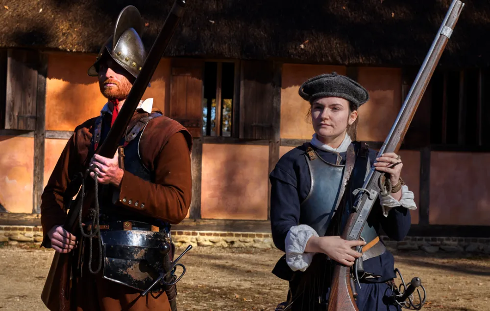 Two soldiers wearing steel breastplates and carrying muskets stand guard outside a thatched house