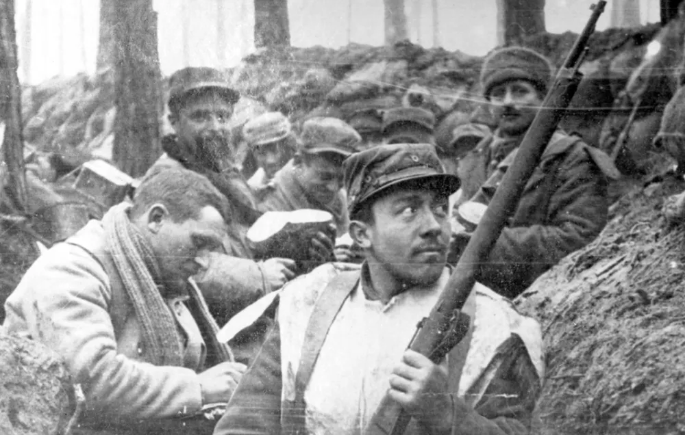 A black and white photograph of a French soldier taking cover in a shallow trench in a wooded area. More soldiers appear behind him, all taking cover.