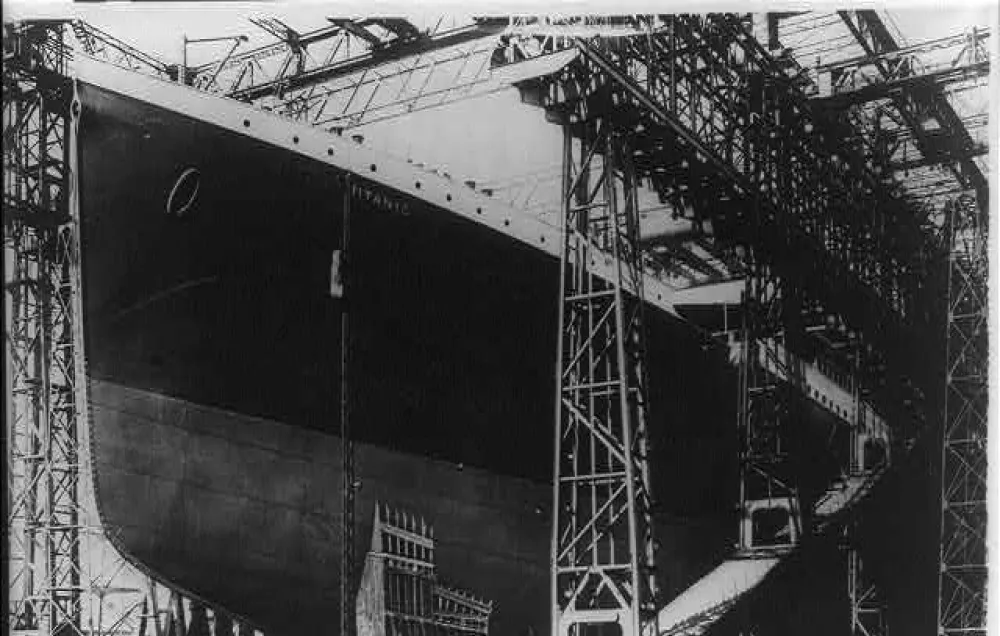 A black and white photograph of an ocean liner in mostly black paint being built while surrounded by scaffolding.