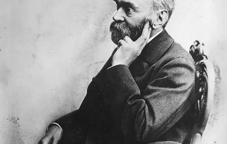 A black and white photograph of a bearded man in profile, seated and leaning one hand against the side of his face.