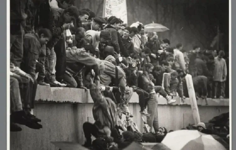A black and white photo of people on top of a concrete wall helping each other up to stand or cross the barrier.