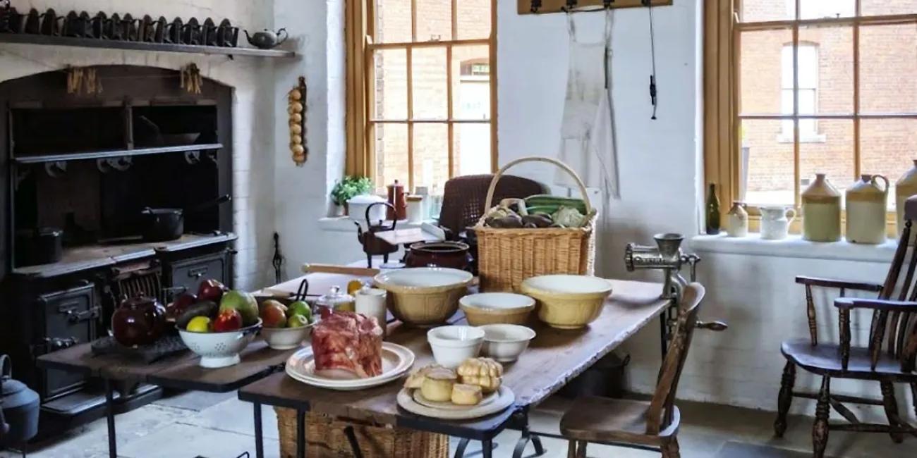 19th century kitchen with 2 large windows a stove, table with bowls and food items and 2 chairs