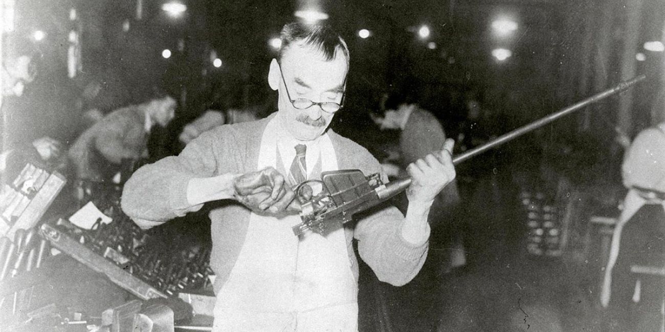 Middle-aged man in a factory wearing glasses inspecting the trigger assembly of a rifle