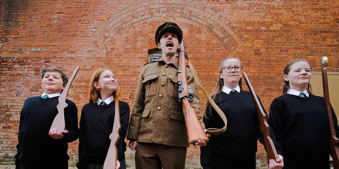 Man in WW1 uniform shouts, four students stand either side of him looking forward, one looks up at him with a bemused expression