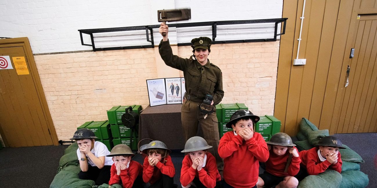 Woman dressed in WW2 ATS Uniform stands with a gas rattle in the air. At her feet are crouched school children wearing tin hats and cupping their hands over their mouth