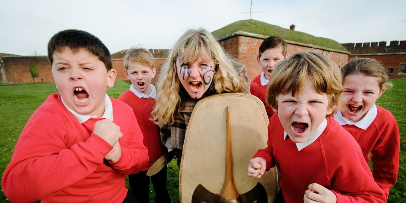 Iron Age Warrior with blue face paint and holding a shield screams at the camera. Around her are school children screaming as well.