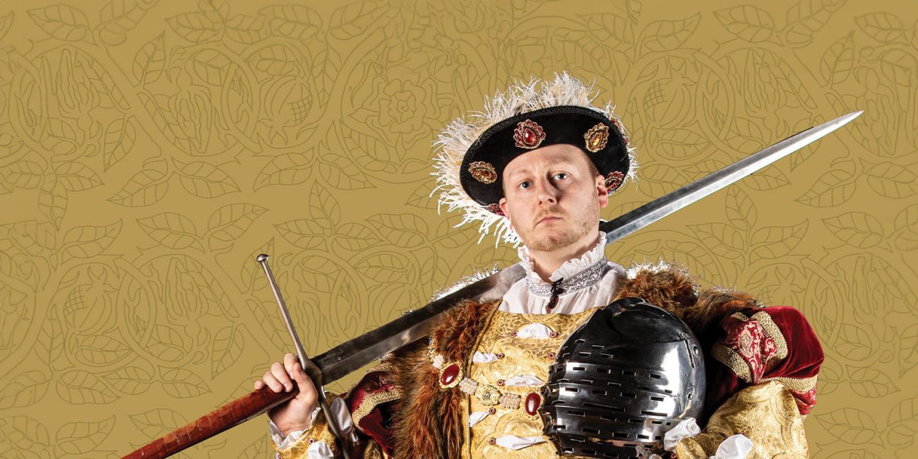 Man dressed in period Tudor costume with sword resting on his right shoulder and a knights helmet in his left hand