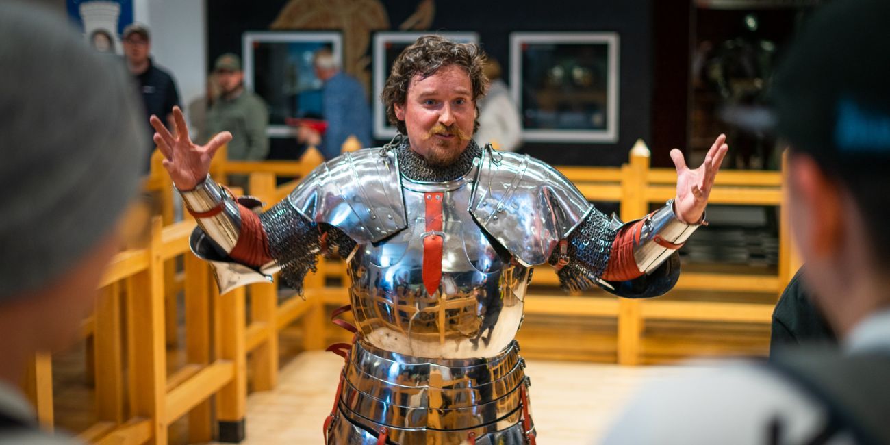 a man dressed in shining armour addresses the audience excitedly with his arms spread wide