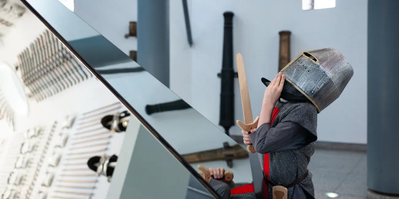 child dressed as a knight gazes upwards in front of a mirror reflecting the interior of the hall of steel
