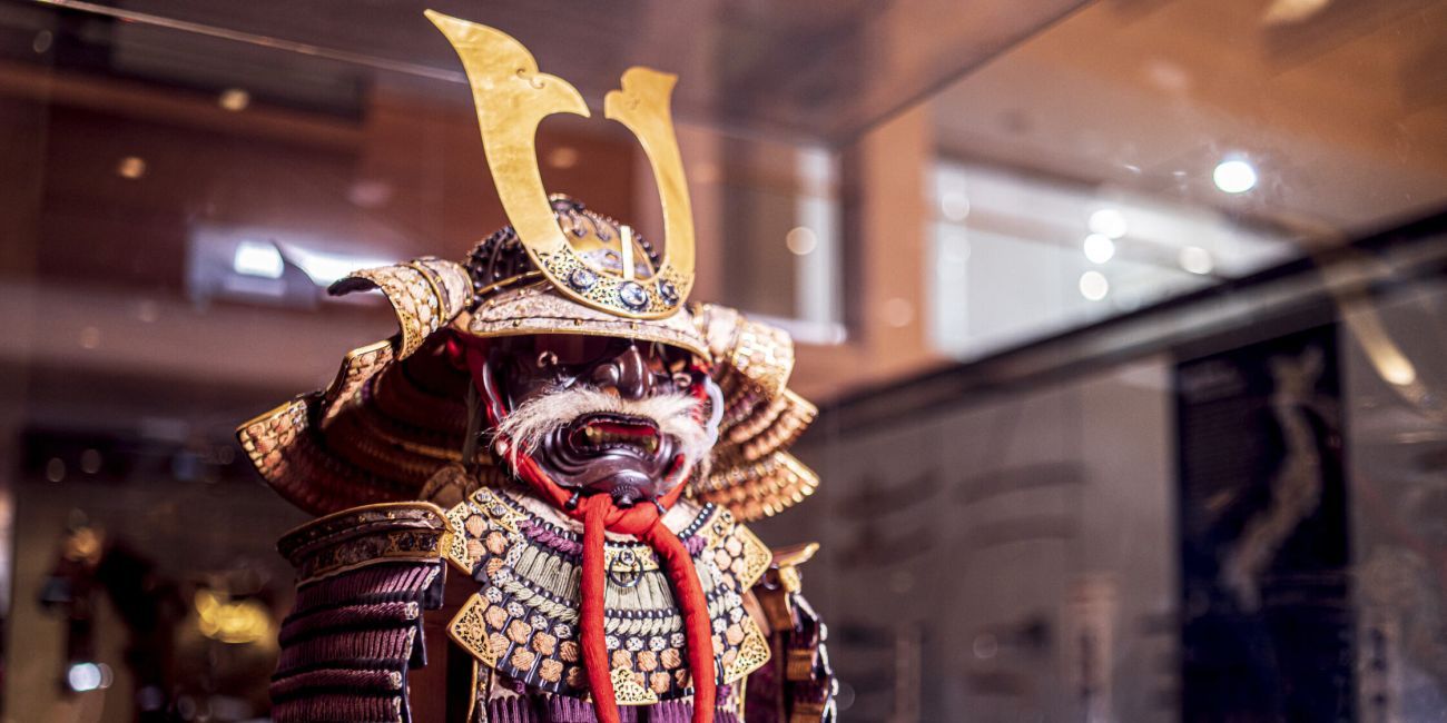 a purple and gold Samurai armour with a fearsome mask in the form of a demon with white whiskers stands guard in the gallery