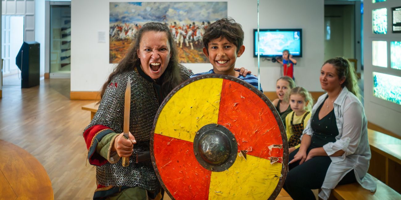 a fearsome Viking shield maiden is poised for action, a boy smiles nervously beside her behind a large round yellow and red shield
