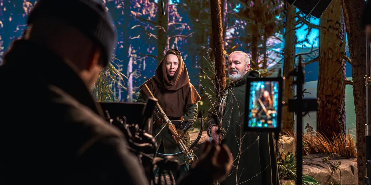 Male and female in hooded clothing being filmed for promotional content with digital woodland scene behind.