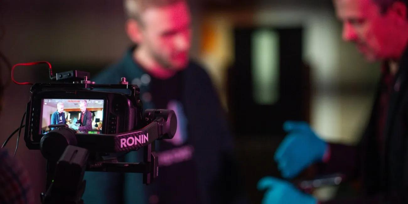 Behind the scenes filming, showing rear camera screen with two History Hit presenters in the background. 