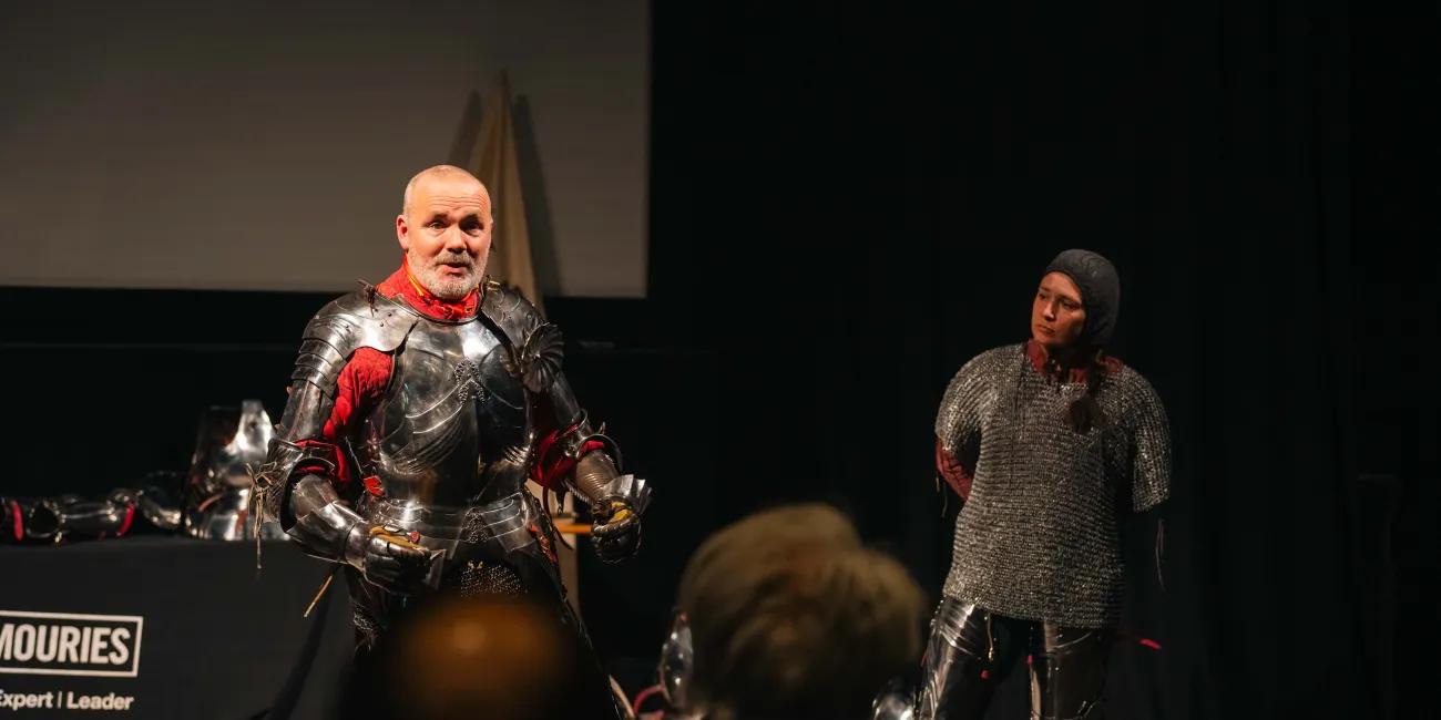 Man in armour giving a demonstration to the audience with female wearing chainmail beside him.