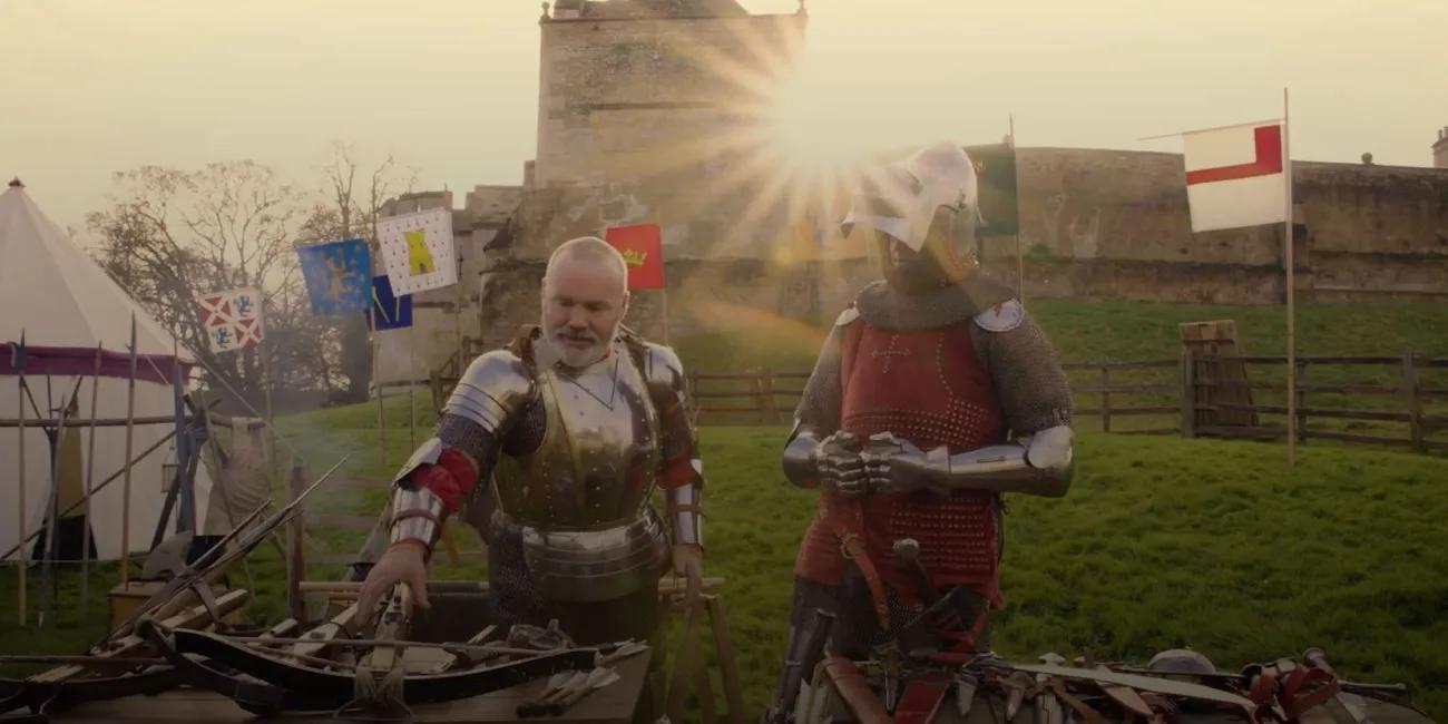 Image of two men in armour looking at weapons on a table with sun setting behind a castle in the background.