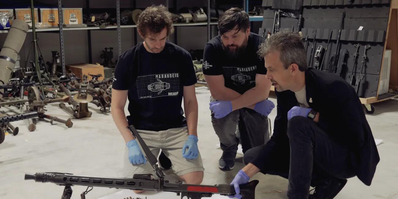 Jonathan Ferguson talking to two visitors whilst holding firearm and also other firearms in the background