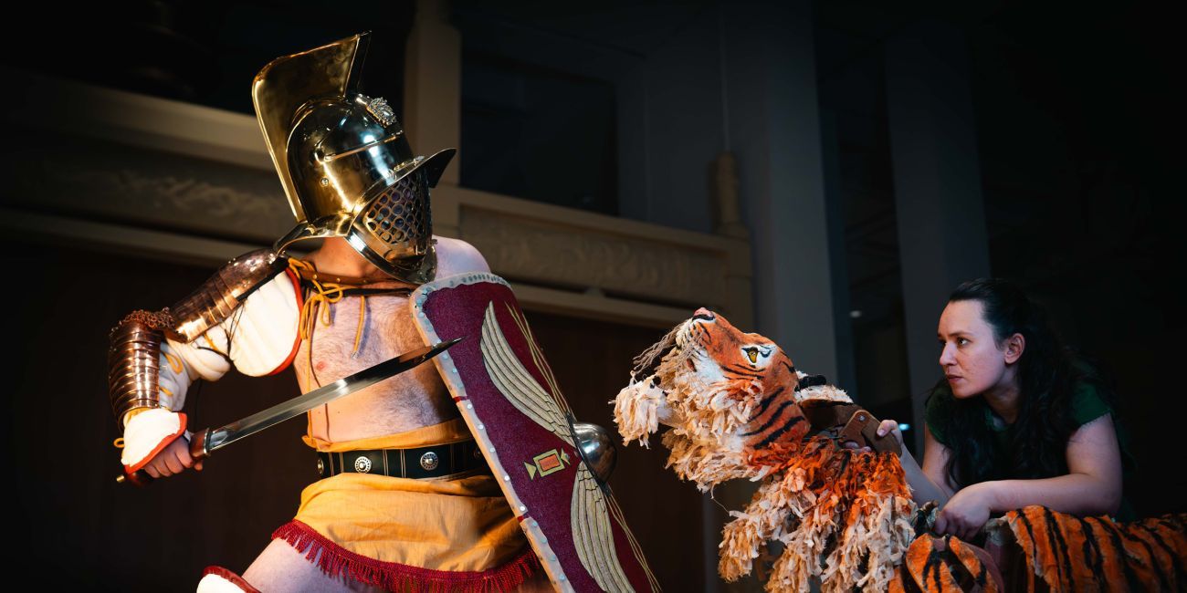 Gladiator in traditional armour with sword and shield confronts a large tiger puppet operated by a person