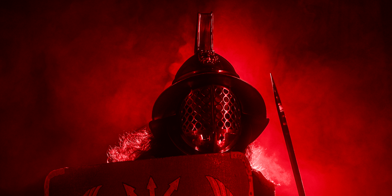 A person in Roman gladiator armour holds a shield and sword, standing in dramatic red lighting and smoke, creating a powerful and intense atmosphere