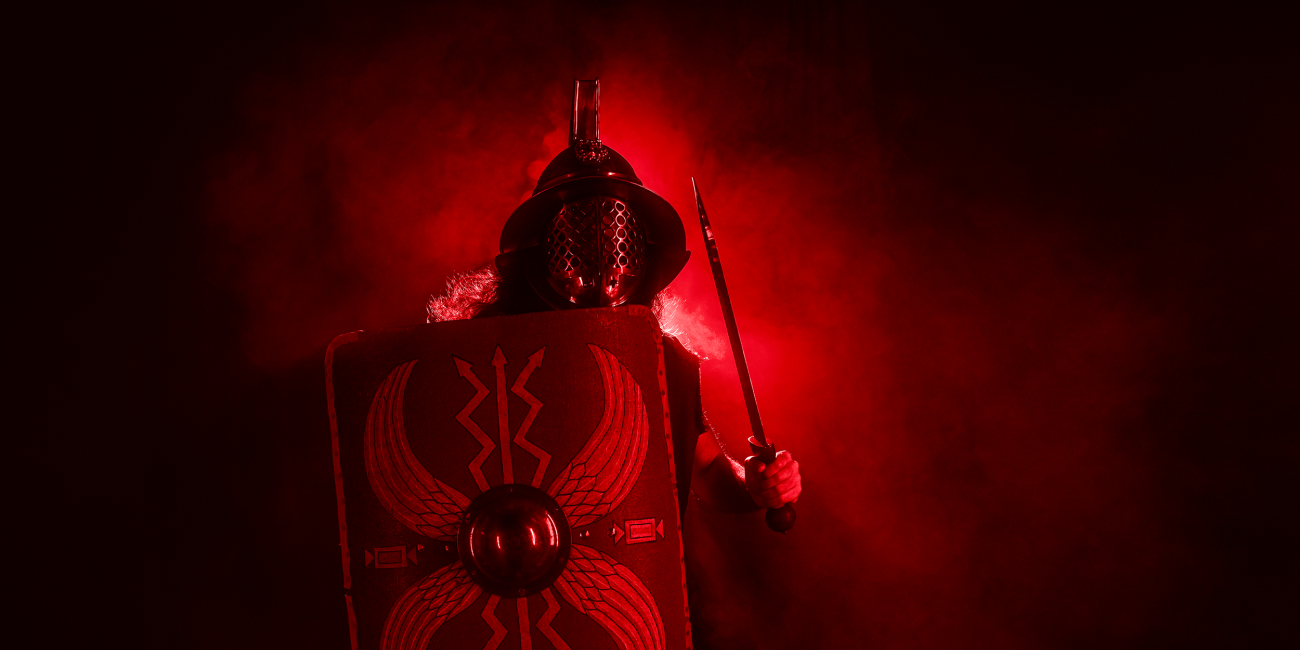 A person in Roman gladiator armour holds a shield and sword, standing in dramatic red lighting and smoke, creating a powerful and intense atmosphere