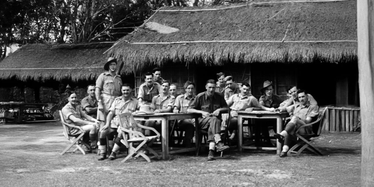 Group of soldiers seated and standing around two wooden tables on grass near a thatched-roof building, with trees in the background