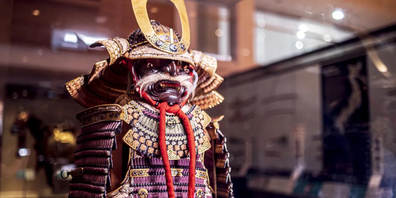 a purple and gold Samurai armour with a fearsome mask in the form of a demon with white whiskers stands guard in the gallery