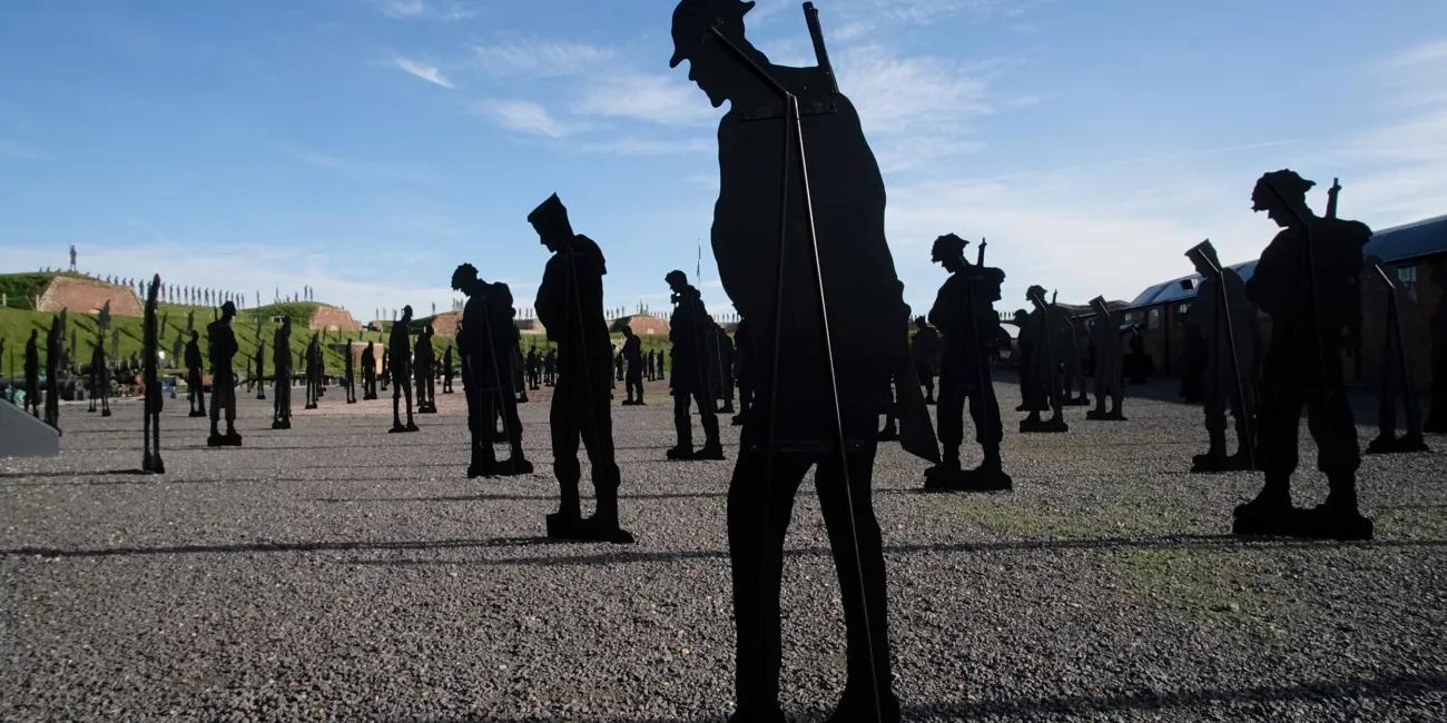 Life-sized soldier silhouettes positioned on a stony ground, heads bowed, evoking remembrance against a bright sky backdrop