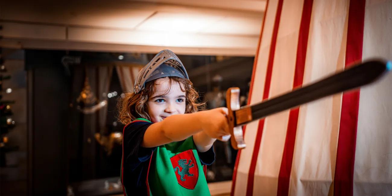A child dressed in medieval-style costume wearing a metal-look helmet and holding a long toy sword outward, standing beside a canvas structure with red vertical stripes.
