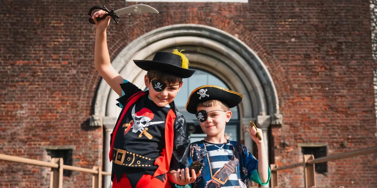 Two young children dressed as pirates pose with raised swords on a bridge, with a large brick entrance behind them displaying the text “FORT NELSON”