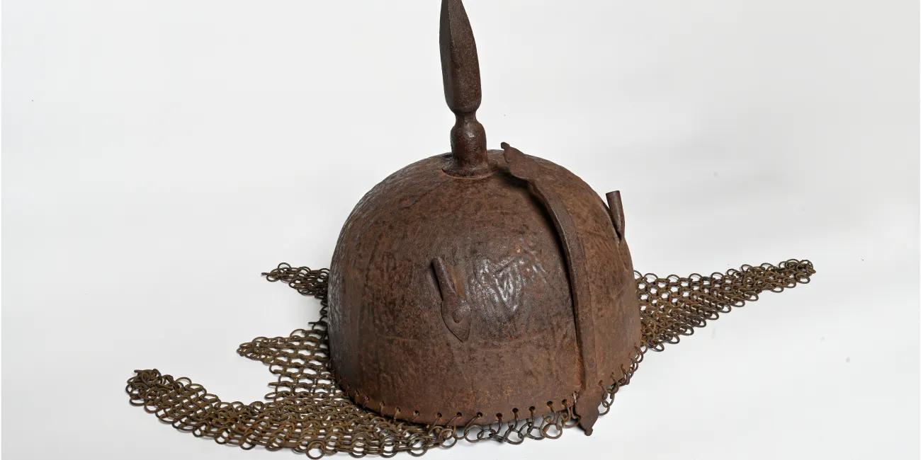 Historical metal helmet featuring a rounded top with a pointed finial and a chainmail neck guard extending outward on both sides
