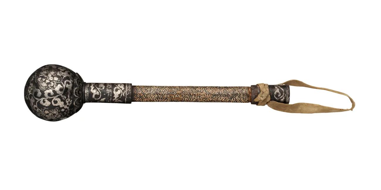 Historical Mongolian mace with a patterned metal ball head, elongated grip wrapped in textured material, and a leather carrying strap, isolated against a white background