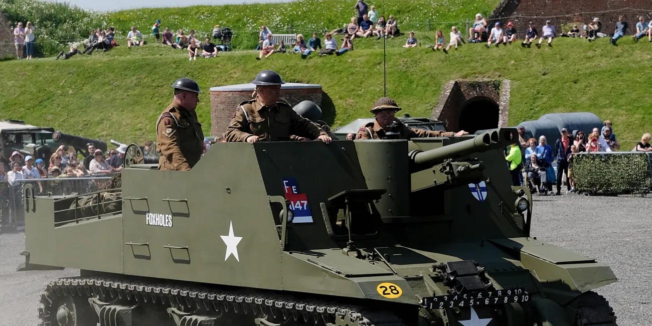 Olive-green self-propelled artillery vehicle with white star markings, moving on gravel with people aboard and a crowd behind