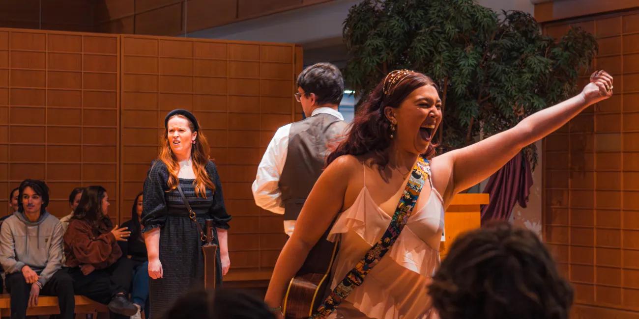 Performer in light dress with guitar strap raises arm while singing on small stage; other cast members and seated audience visible