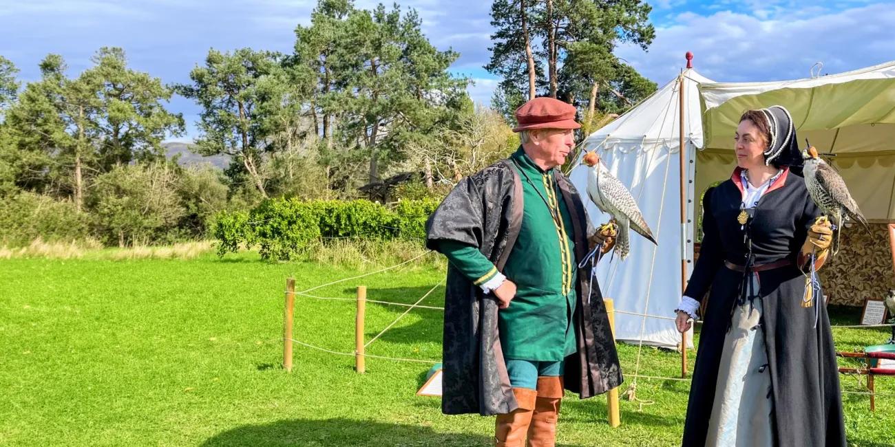 Falconry display outdoors: two costumed people with hooded falcons stand on grass near a white tent and woodland