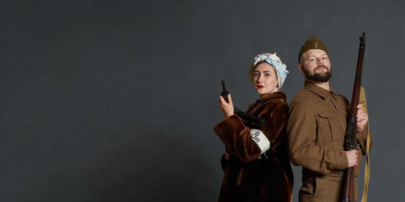 A Second World War female SOE operative and soldier standing back to back looking forward.  The SOE is in civilian dress with a fur coat and head scarf. The soldier is in uniform. Both are holding firearms. 