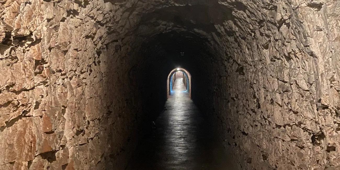 Narrow rock tunnel with rough stone walls and shallow water on the floor, illuminated by light at the far end