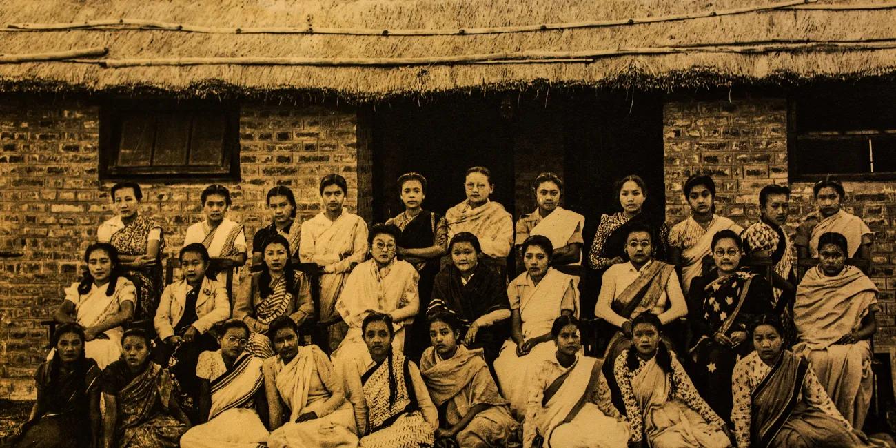 Sepia-toned group of women in saris and uniforms, seated and standing in three rows in front of a brick structure