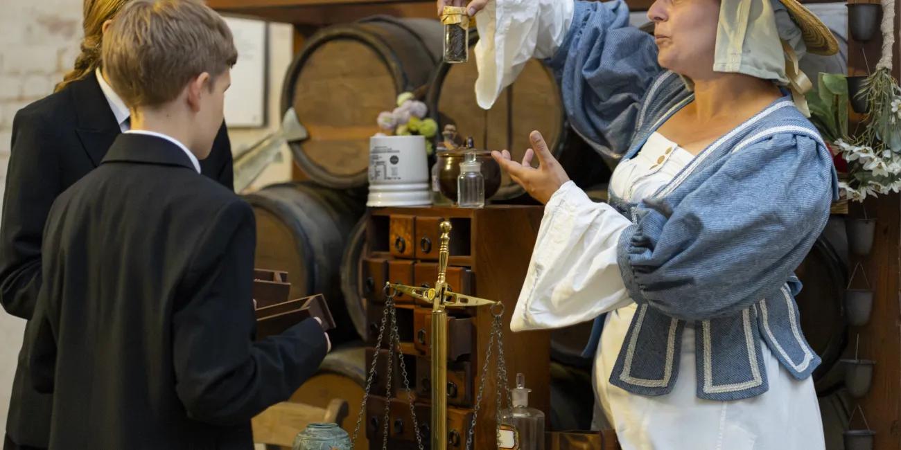 17th-century herbal women displays medicine jar to two school pupils