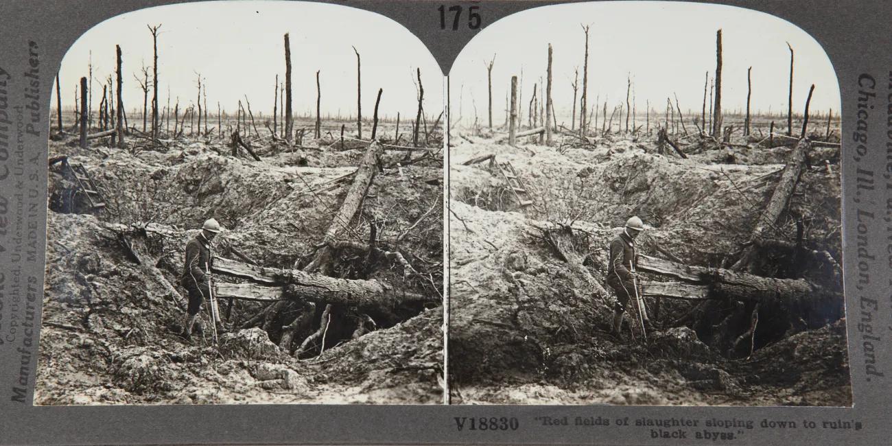 Black-and-white stereoview of a battlefield with shattered tree trunks, deep craters and a soldier standing in foreground