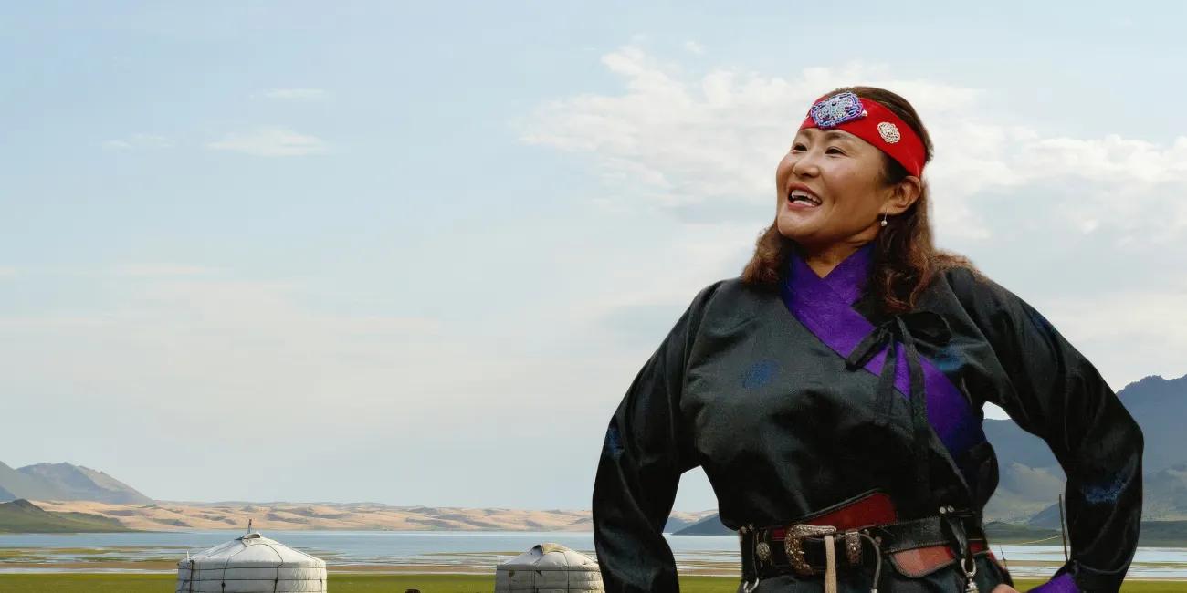 Figure in black attire with purple trim overlooking open plains, yurts and a calm lake under a bright sky