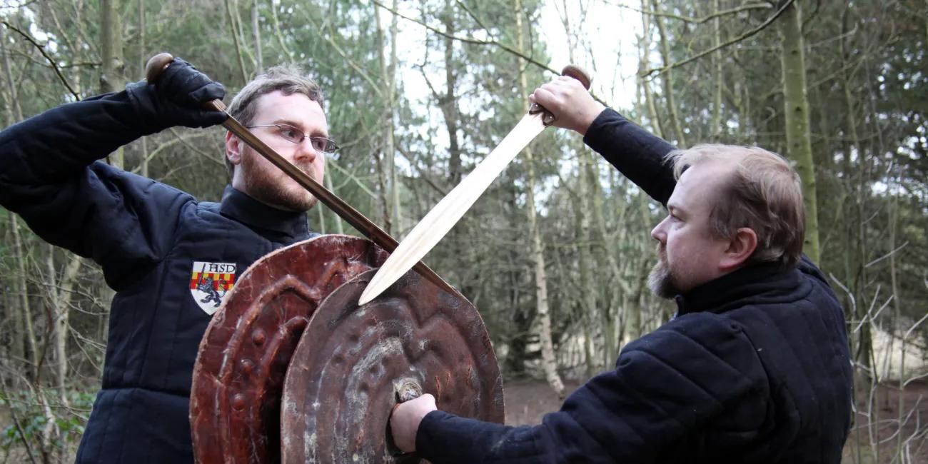 Historical combat reenactment showing crossed swords and large round shields among trees in a forest