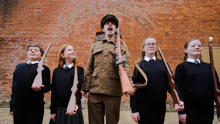 Man in WW1 uniform shouts, four students stand either side of him looking forward, one looks up at him with a bemused expression