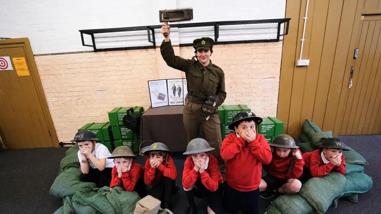 Woman dressed in WW2 ATS Uniform stands with a gas rattle in the air. At her feet are crouched school children wearing tin hats and cupping their hands over their mouth