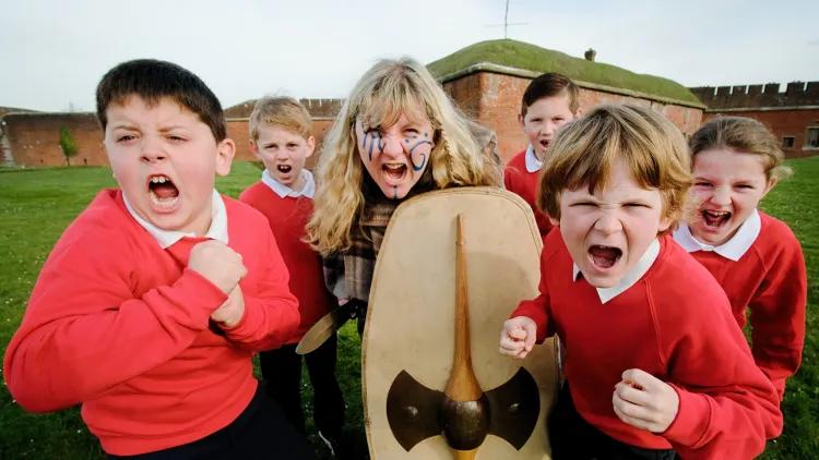 Iron Age Warrior with blue face paint and holding a shield screams at the camera. Around her are school children screaming as well.