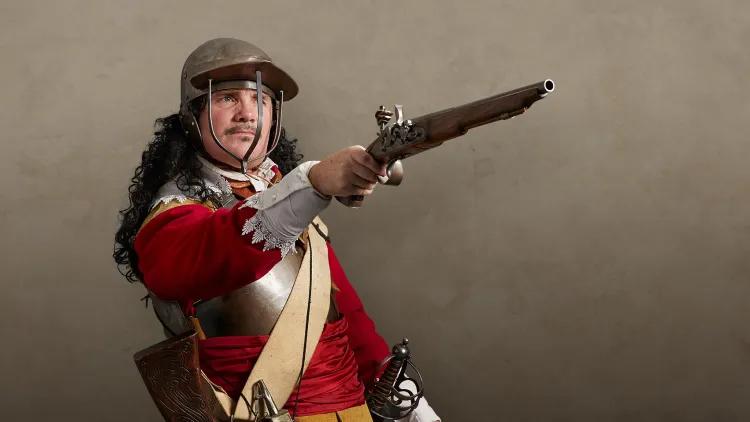 Royalist cavalier soldier dressed in a red tunic, buff coat, breastplate and a three-bar helmet, pointing a flintlock pistol