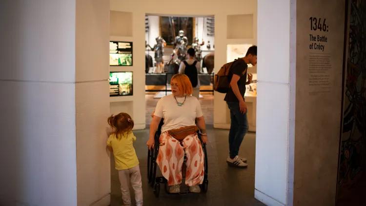 a visitor in a wheelchair navigates a gap in a gallery comfortably while passing a small child who leans against the wall